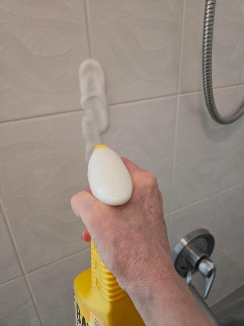 a hand holding a yellow cleaning spray bottle the nozzle is putting foam cleaner on the wall of the shower a woman's hand shown operating the bottle
