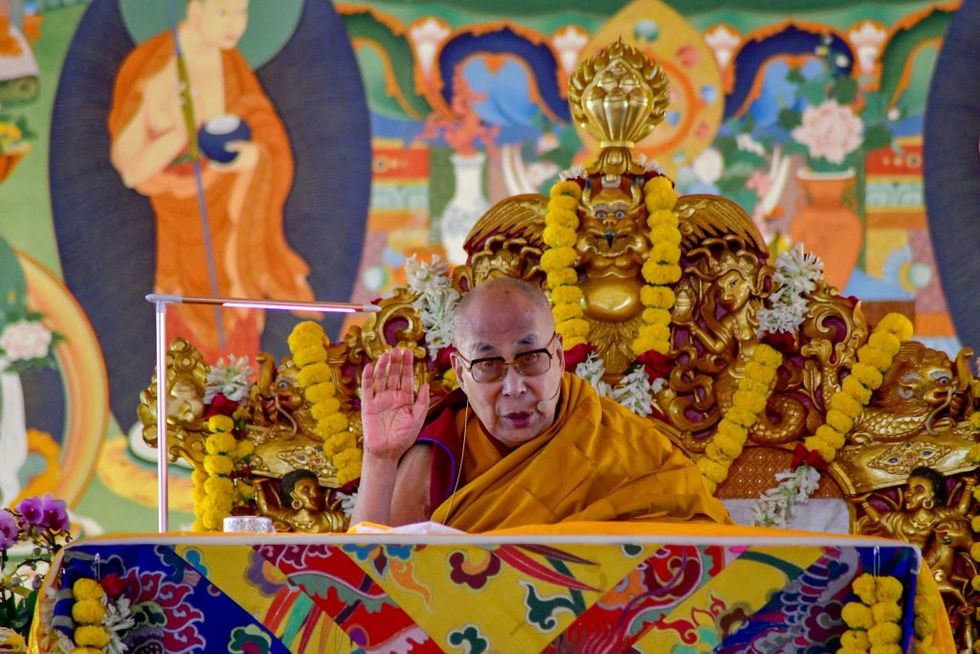 the dalai lama raising his hand while sitting in a decorative chair
