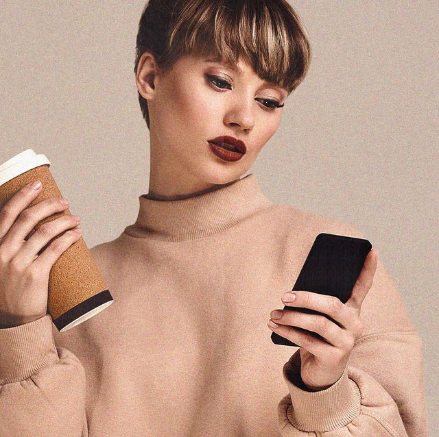 a young woman in a beige jumper in front of a beige background holding a coffee in one hand and a phone in the other