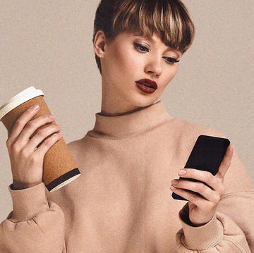 a young woman in a beige jumper in front of a beige background holding a coffee in one hand and a phone in the other