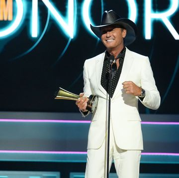 tim mcgraw smiles and stands behind a microphone while on a stage, he wears a white suit with a black collared shirt with white polka dots and a black cowboy hat, he holds a gold award in one hand, behind him is a background that says academy of country music honors