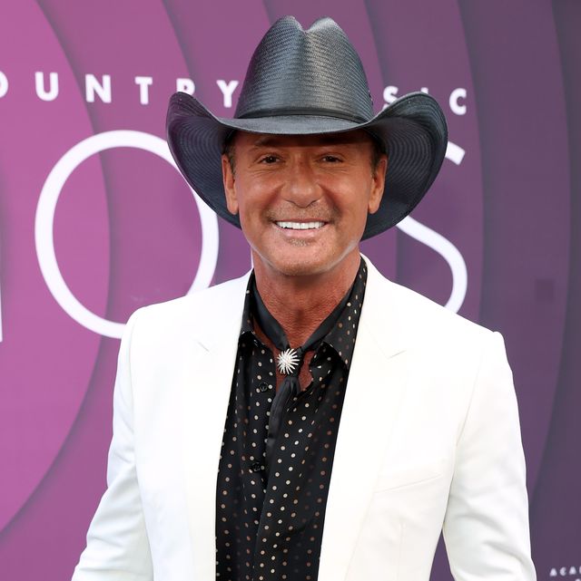 tim mcgraw smiles at the camera while standing in front of a purple background, he wears a white suit jacket, black collared shirt with white polka dots and a black cowboy hat