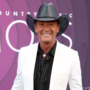 tim mcgraw smiles at the camera while standing in front of a purple background, he wears a white suit jacket, black collared shirt with white polka dots and a black cowboy hat