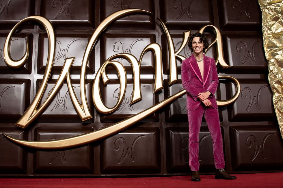 timothée chalamet smiling as he stands in front of a giant chocolate bar with the wonka logo on it