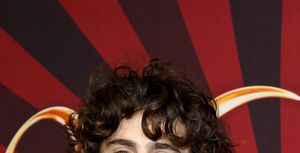 timothee chalamet smiles at the camera, he wears a multicolored plaid suit jacket with a woven necklace, he stands in front of red and brown background
