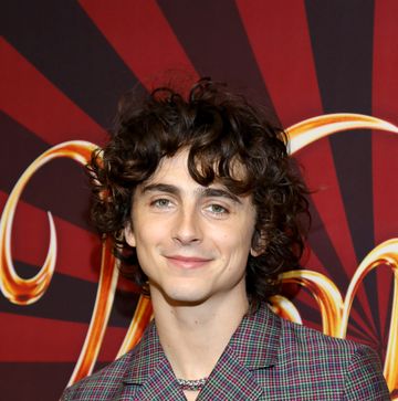 timothee chalamet smiles at the camera, he wears a multicolored plaid suit jacket with a woven necklace, he stands in front of red and brown background