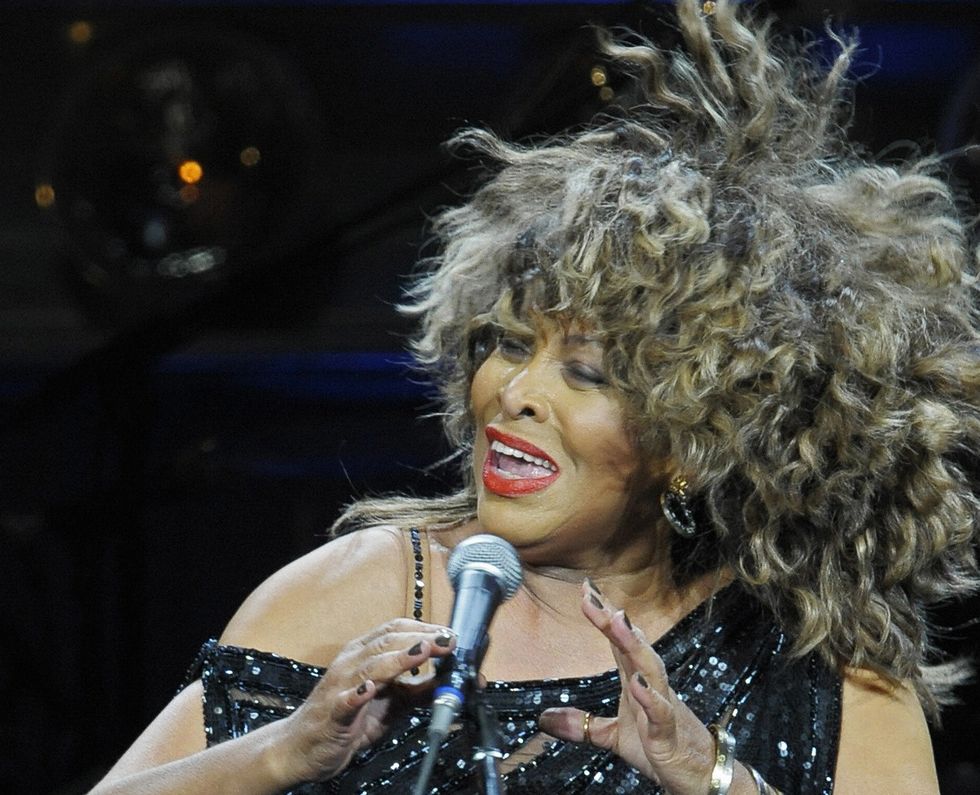 tina turner sings into a microphone with her head cocked to the left, she wears a black sequin top and gold jewelry