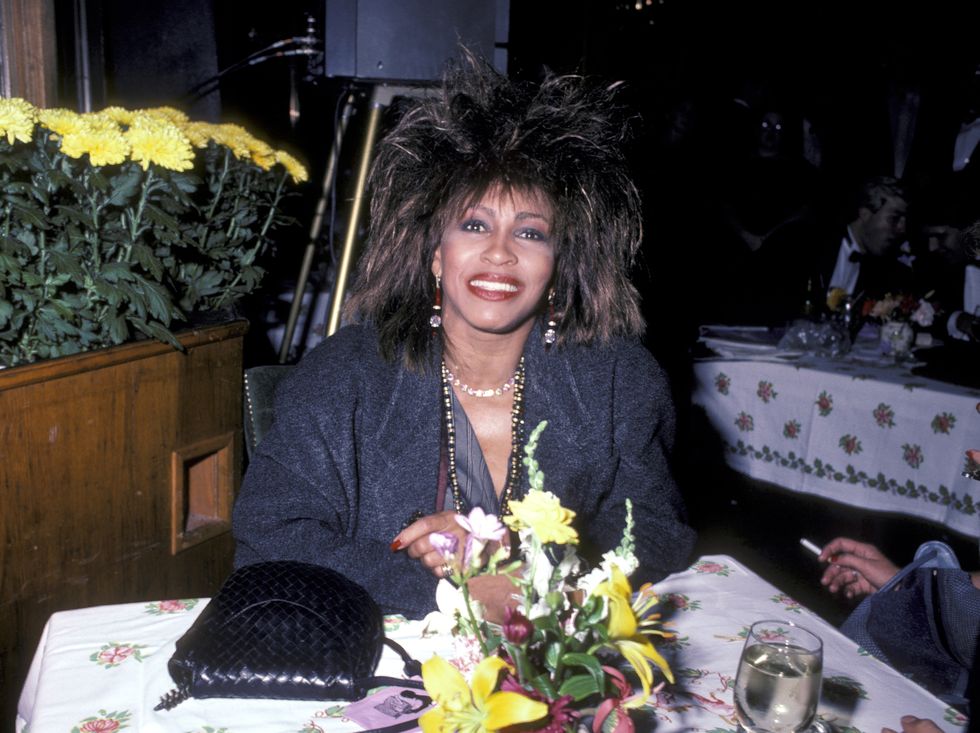 tina turner smiles while seated at a table with a floral arrangement on it