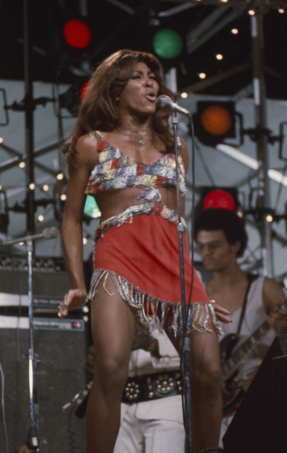 tina turner sings into a microphone on a stand, she wears a revealing minidress with beaded fringe