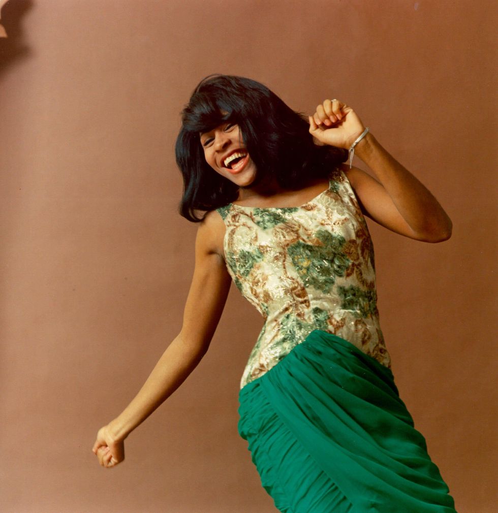 tina turner smiles while posing for a photo, she wears a patterned sleeveless dress with a green skirt