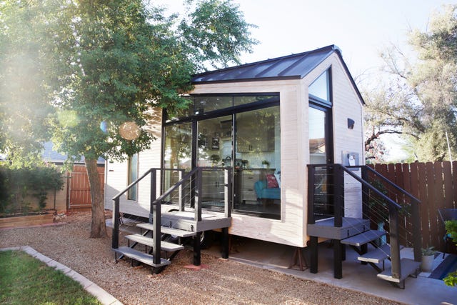 a tiny house with large glass windows, sits in the backyard, surrounded by a wooden fence and trees