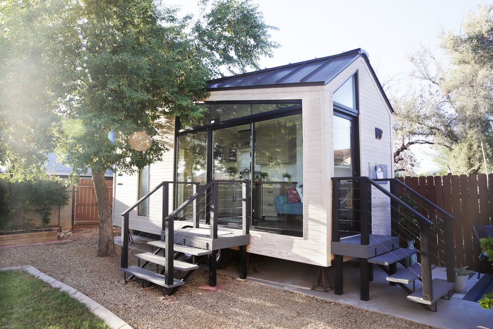 a tiny house with large glass windows, sits in the backyard, surrounded by a wooden fence and trees.