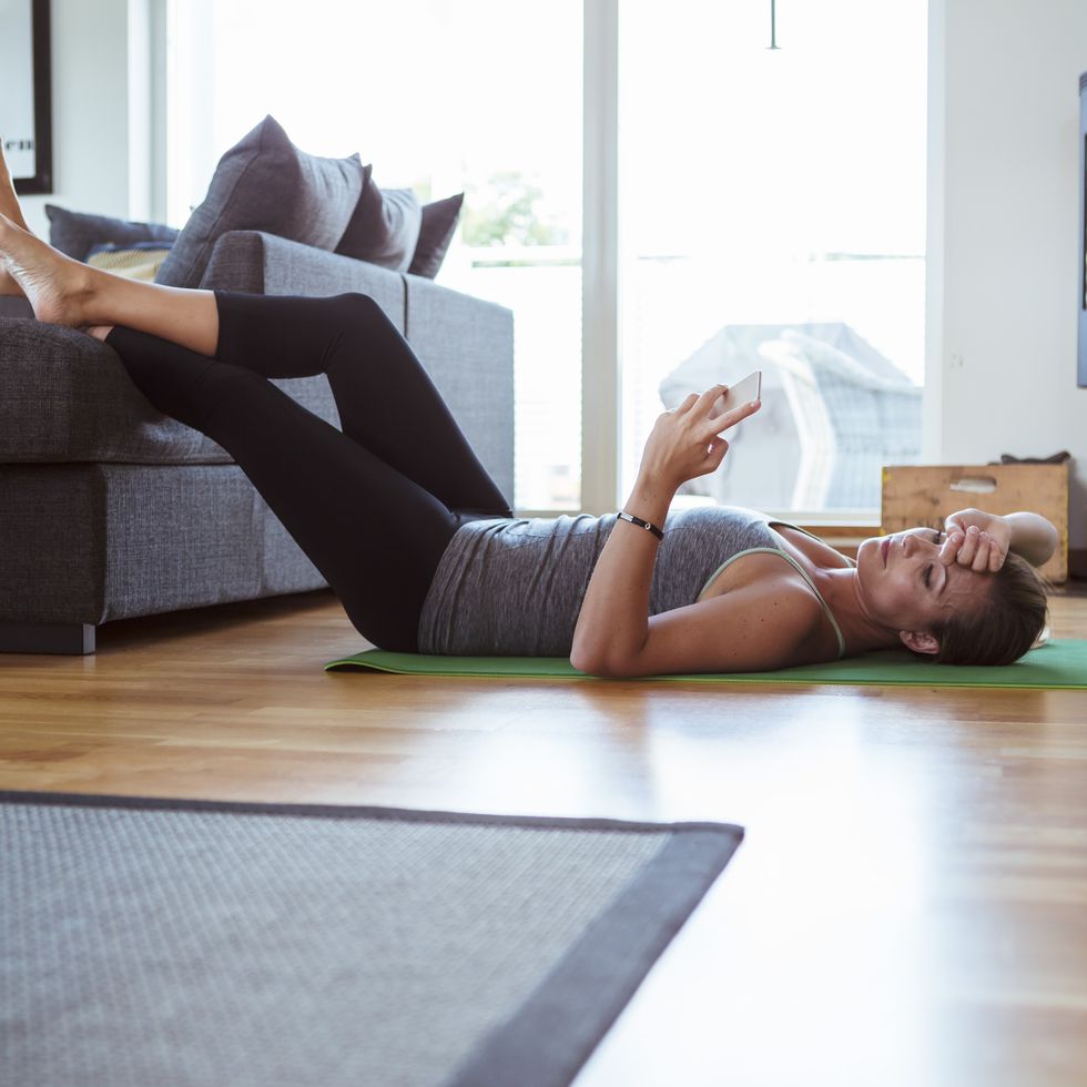 tired woman using mobile phone while lying on exercise mat in living room