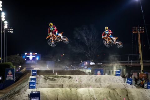 two dirt bike racers fly in the air over an obstacle in the sand section