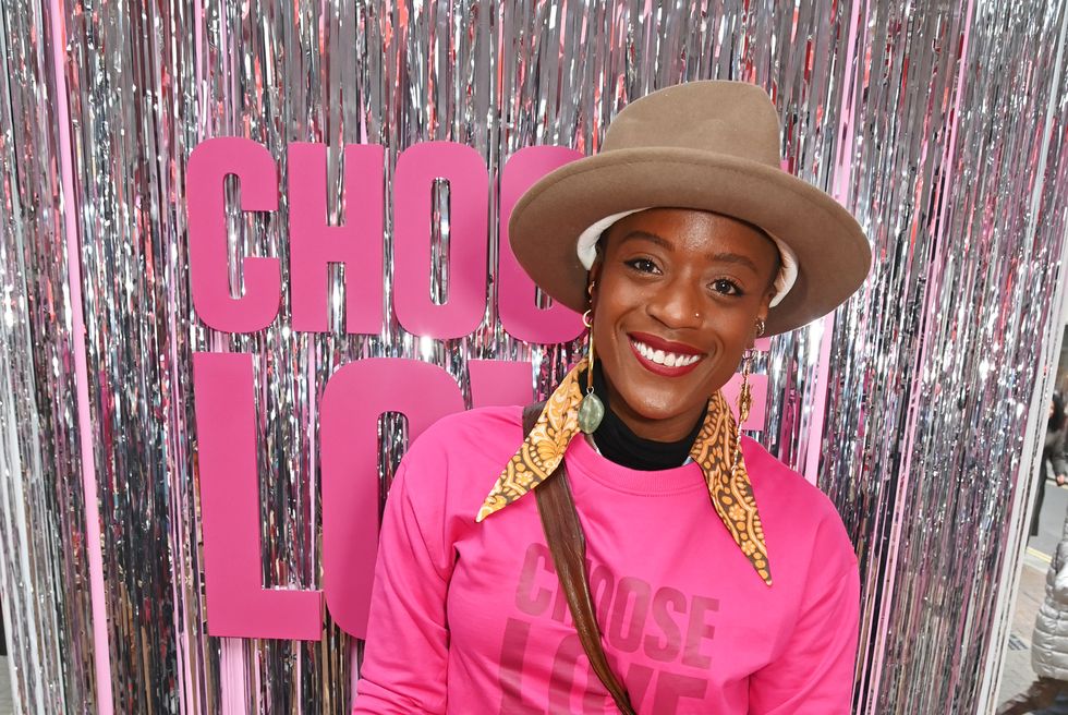 tnia miller, a woman wearing a bright pink sweater and a large fedora hat, standing in front of a wall of silver streamers and text that reads choose love, smiling for the camera