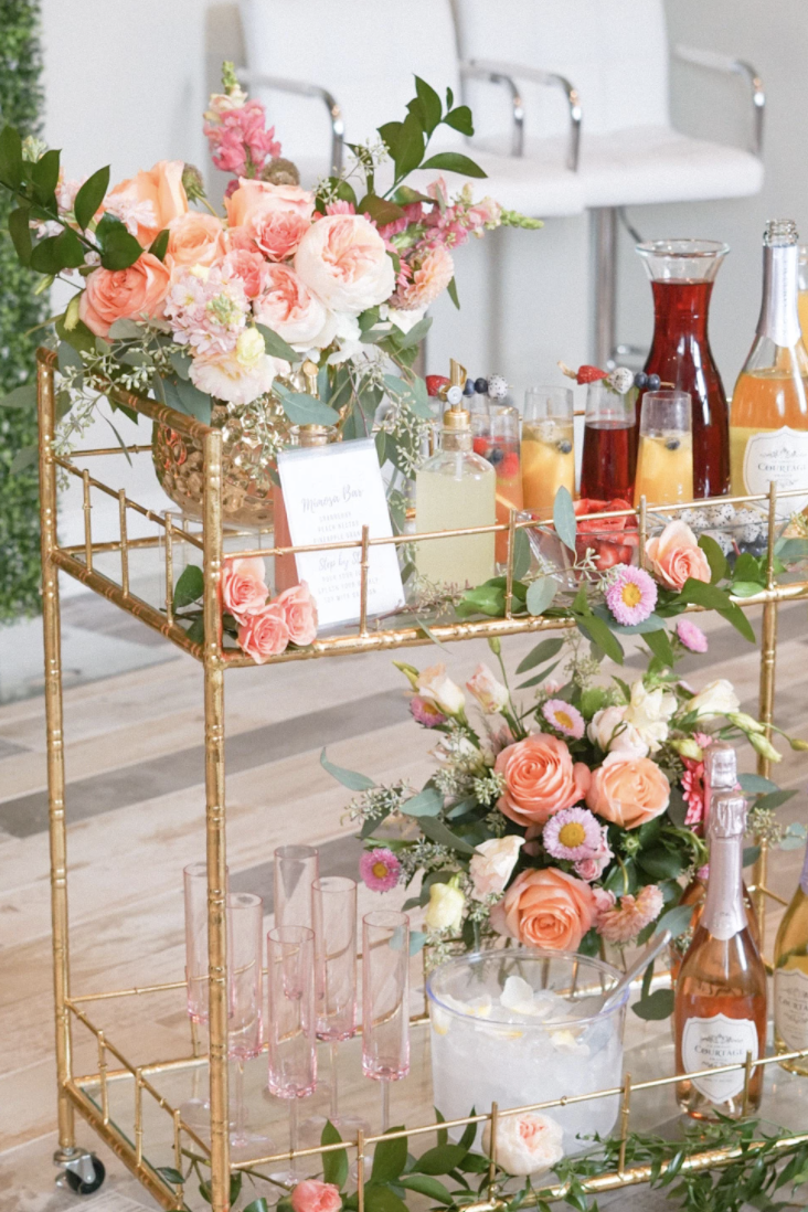 gold rectangle bar cart with coral roses and greenery on it along with juice, champagne, an ice bucket, and pink glasses