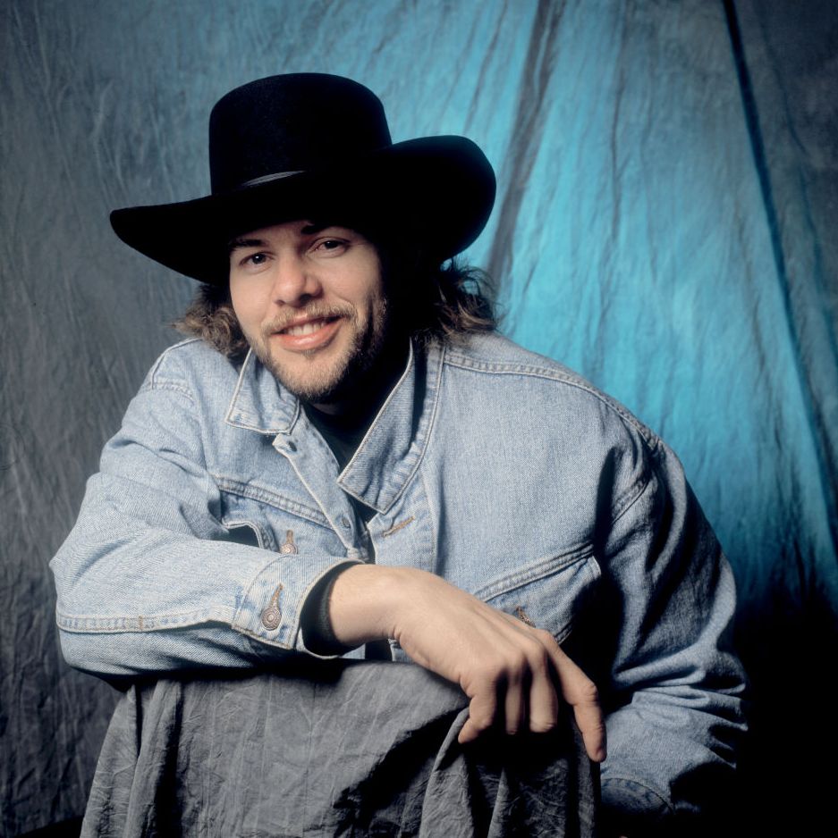 toby keith smiles at the camera while sitting in front of a background lit with a blue light, he wears a light colored jean jacket and a black cowboy hat