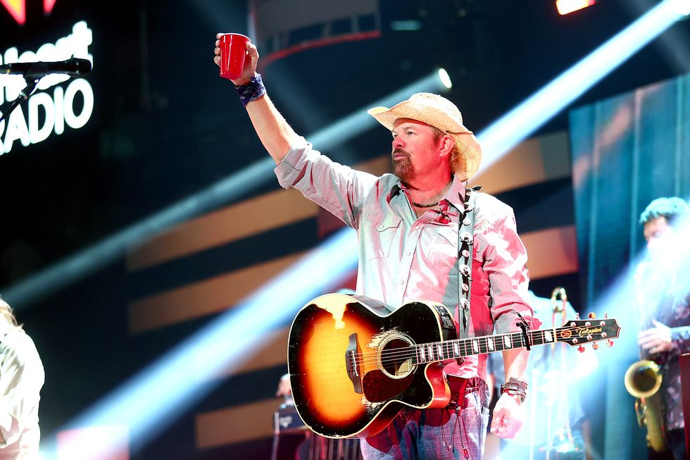toby keith stands on a stage with a few spotlights shining down, he holds a red solo cup above his head and has an acoustic guitar on a shoulder strap, he wears a straw colored cowboy hat, gray collared shirt and jeans