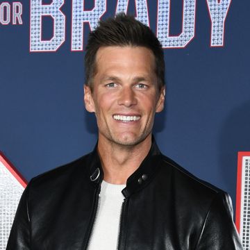 tom brady smiles at the camera, he wears a black leather jacket over a white shirt and stands in front of a navy background