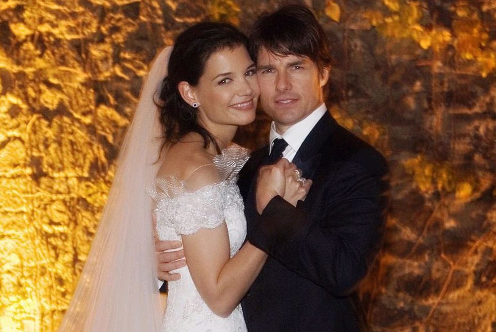 katie holmes and tom cruise smile for a wedding portrait while standing cheek to cheek, she wears a white off the shoulder gown with a veil, he wears a black suit