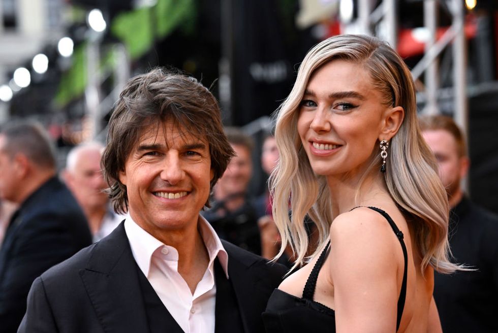 tom cruise, wearing a black suit jacket and pink shirt, smiling alongside vanessa kirby, who wears a black dress