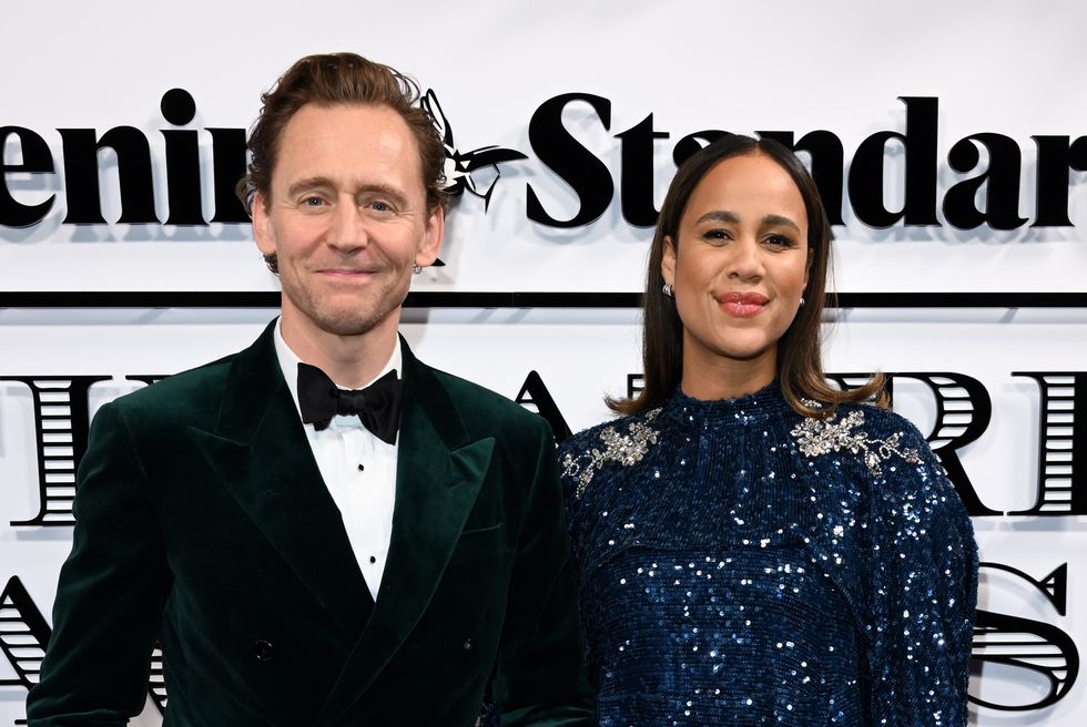 Tom Hiddleston y Zawe Ashton, un hombre y una mujer sonriendo a la cámara tomados de la mano, él viste un traje negro con pajarita y ella viste un vestido largo de lentejuelas azules.