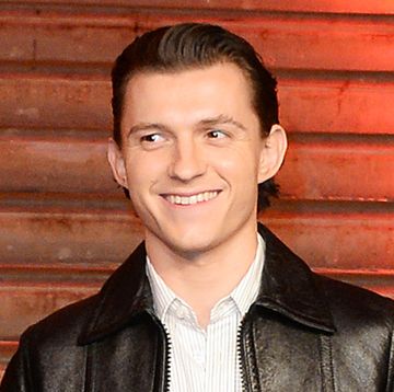 tom holland, standing on some steps, smiles at cameras during a spider man press event