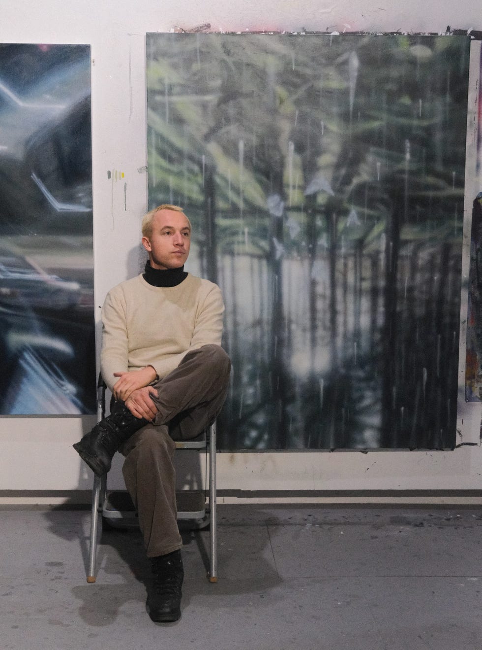 person sitting on a metal folding chair in an art studio setting with abstract artwork on the walls