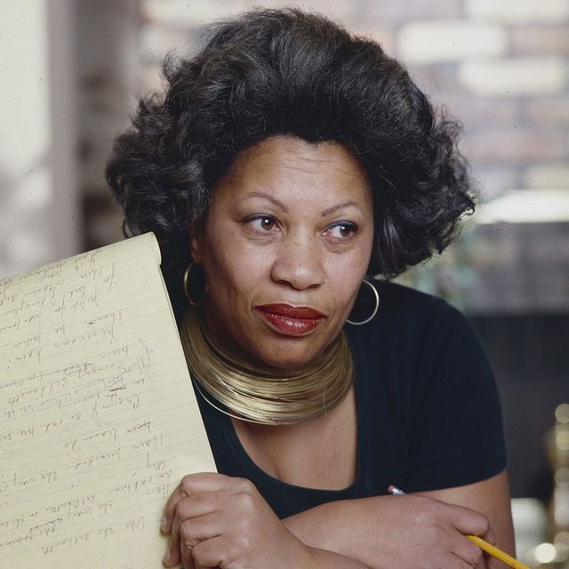 toni morrison looks to the right of the camera with a neutral expression, she holds a pencil in one hand and a yellow legal pad with notes written on it in the other, her arms are crossed, she wears large hoop earrings, a gold metal necklace and a black t shirt