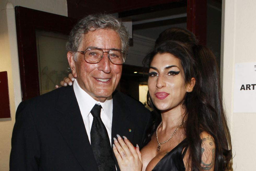 tony bennett and amy winehouse stand next to each other, smile, and pose for a photo, bennett wears a black suit and tie with large glasses, winehouse wears a low cut black dress and has one hand on bennetts chest