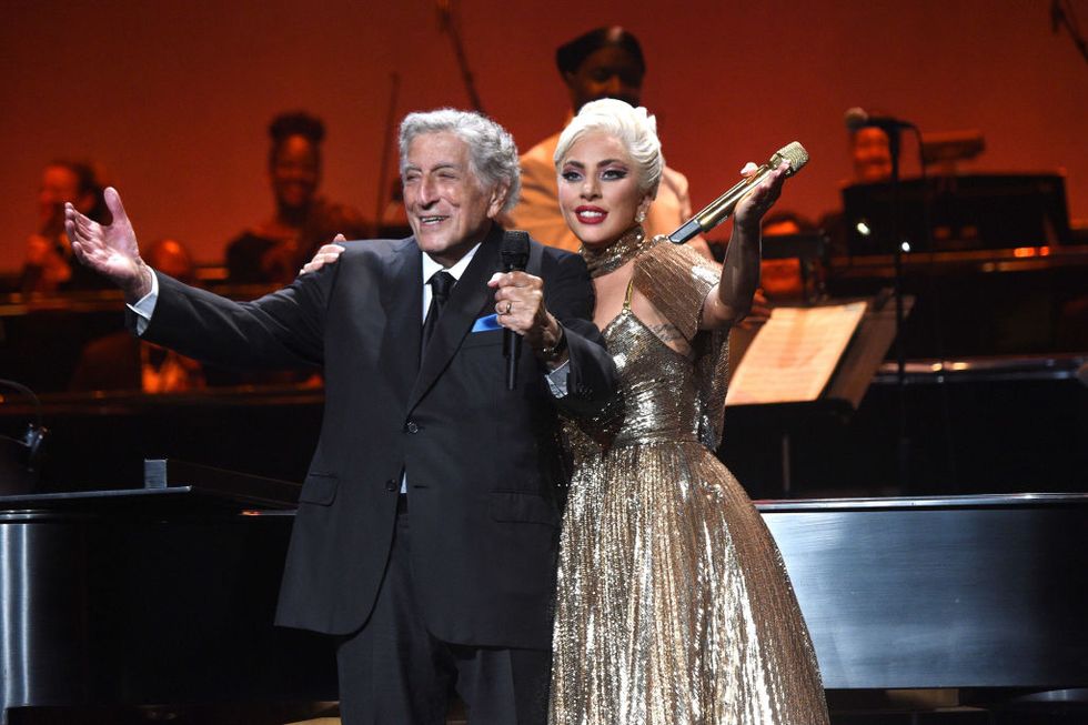 tony bennett and lady gaga stand onstage and extend their arms to the crowd, bennett wears a black suit and tie with a blue pocket square, lady gaga wears a shimmering bronze gown and holds a microphone