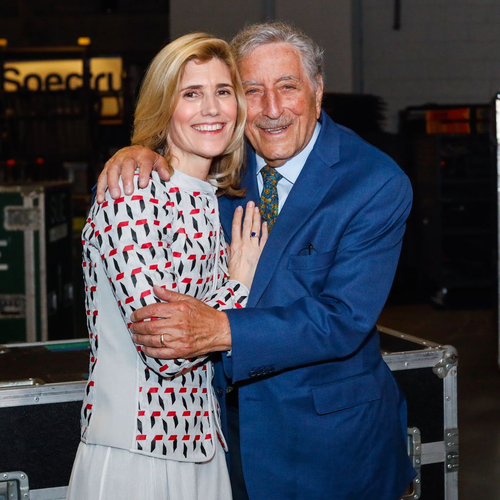 susan benedetto and tony bennett embrace and smile at the camera, benedetto wears a gray jacket with a black, white and red pattern, bennett wears a bright blue suit with a patterned tie