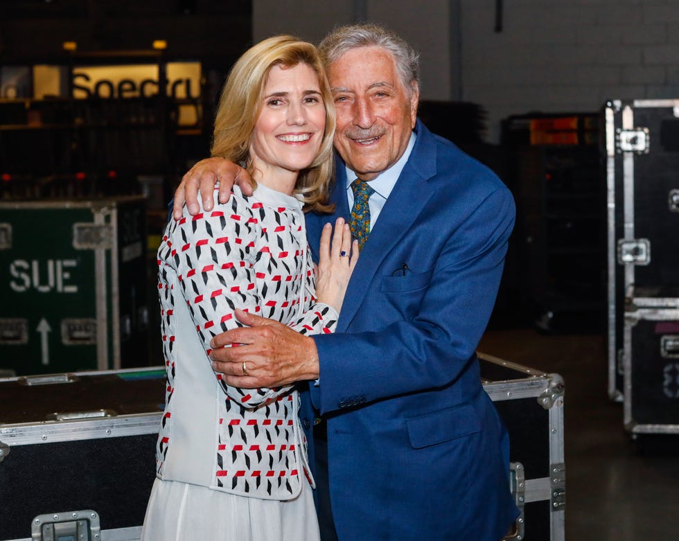 susan benedetto and tony bennett embrace and smile at the camera, benedetto wears a gray jacket with a black, white and red pattern, bennett wears a bright blue suit with a patterned tie