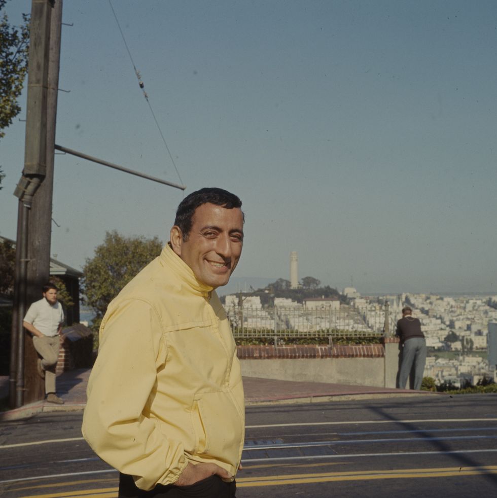tony bennett smiles at the camera while walking on a street with his hands in his pants pockets, he wears a yellow jacket and black pants, behind him is a view of coit tower and surrounding buildings in san francisco