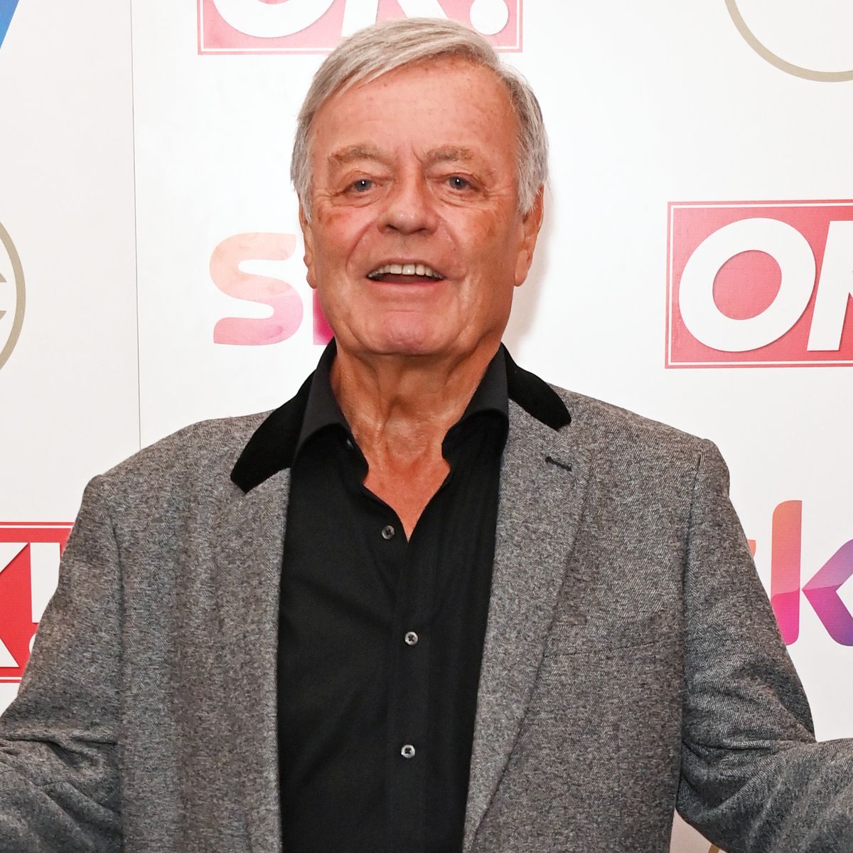 tony blackburn, an older man stands looking at the camera with his hands outstretched, wearing black shirt and trousers with grey jacket