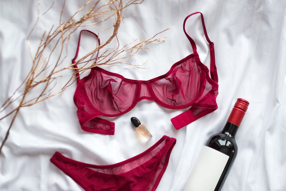 Top view fashion red lace lingerie with bootle of red wine and perfume. Set of woman essential accessory and underwear. Flat lay on white bedsheets background. Shopping and fashion concept.