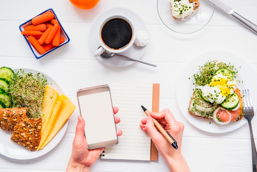 top view female hands holding smart phone and writing in notebook on served white wooden table with breakfast dishes day diet planning and healthy eating concept selective focus, copy space