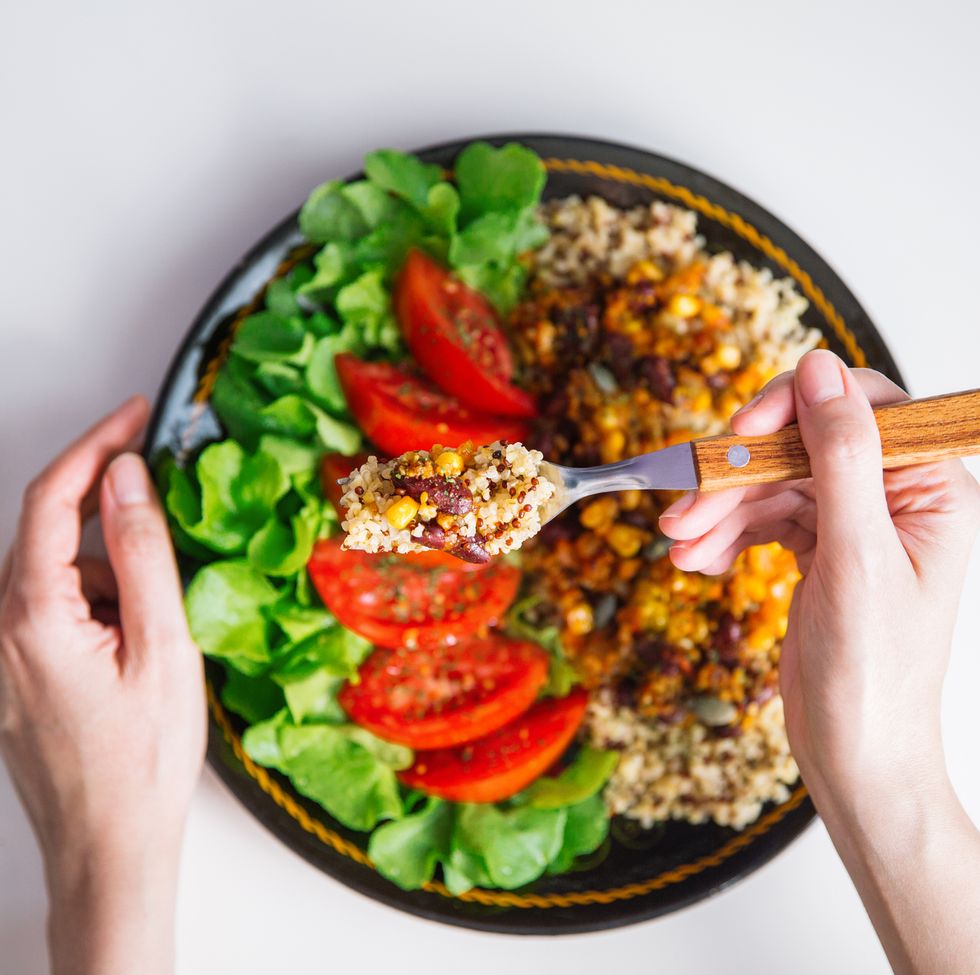top view of female hands at dinner table holding fork above plate with quinoa, red beans, corn, tomato and green salad