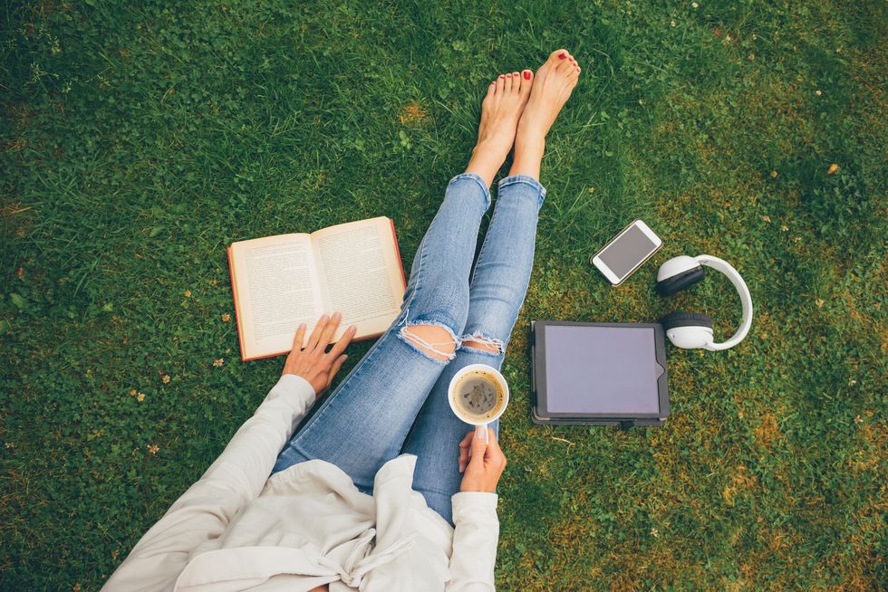 top view of woman sitting in park garden on the green grass with smartphone, headphones, tablet, book and coffee in the hand