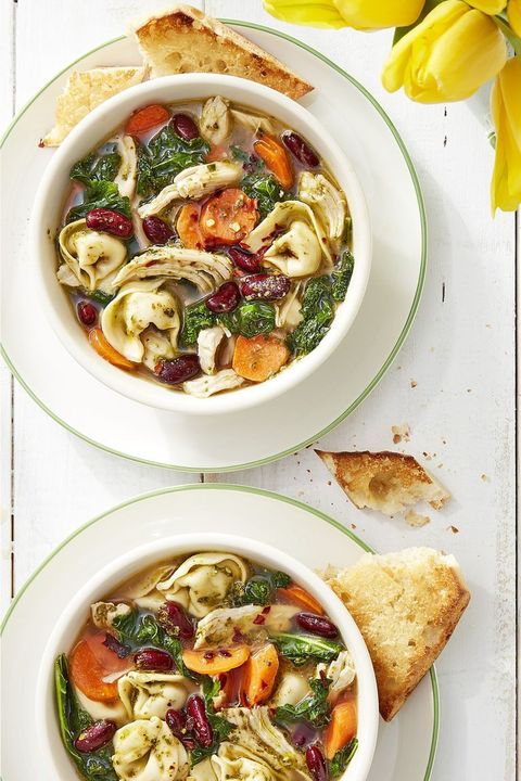 2 bowls of colorful minestrone soup filled with tortellini pasta, carrots, kidney beans, shredded chicken, kale, and pesto, served with crusty toasted bread