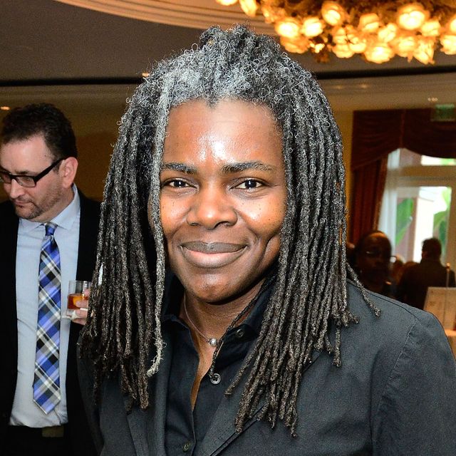 tracy chapman smiles at the camera while standing inside an event space with a chandelier, she wears a black jacket and black collared shirt, her dreads are slightly gray at the roots and reach past her shoulders
