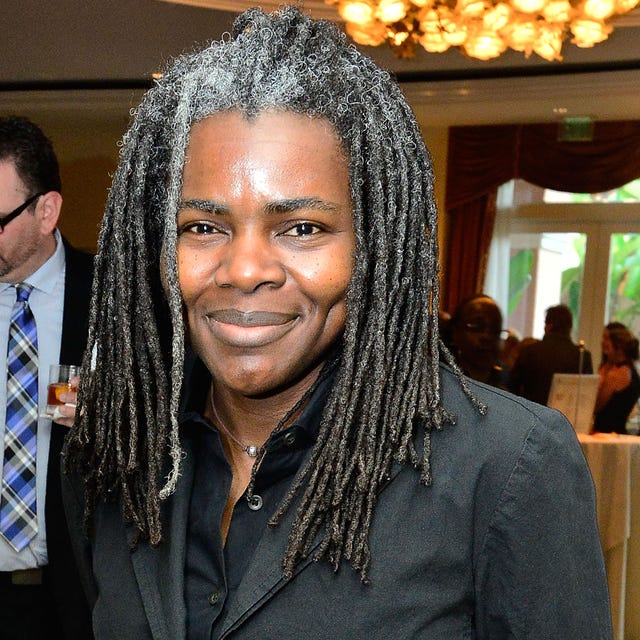 tracy chapman smiles at the camera while standing inside an event space with a chandelier, she wears a black jacket and black collared shirt, her dreads are slightly gray at the roots and reach past her shoulders