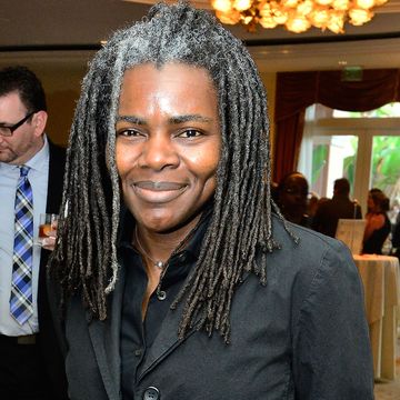 tracy chapman smiles at the camera while standing inside an event space with a chandelier, she wears a black jacket and black collared shirt, her dreads are slightly gray at the roots and reach past her shoulders
