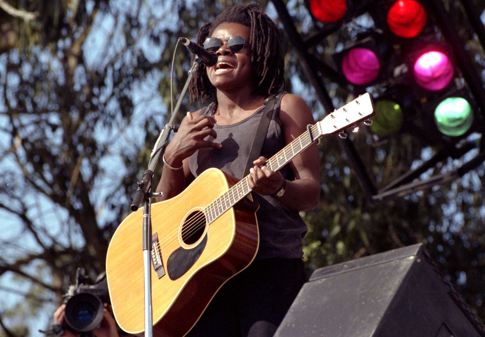 tracy chapman, wearing a black dress and sunglasses, stands on an outdoor stage and sings into a microphone while holding an acoustic guitar