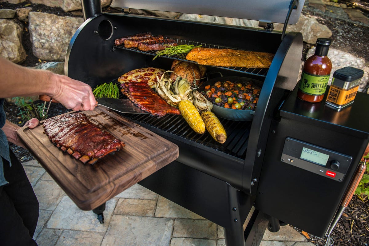 someone taking ribs off of a traeger grill while pizza, steaks, salmon, and vegetables are cooking