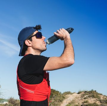 a trail runner with cap and sunglasses drinking in a bottle against a blue sky.