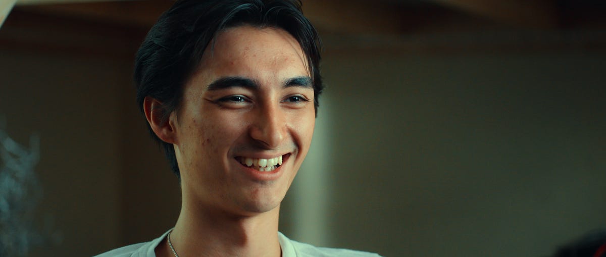 a young man smiles as he looks to the right, only part of his light colored shirt is visible cr. courtesy of netflix © 2025