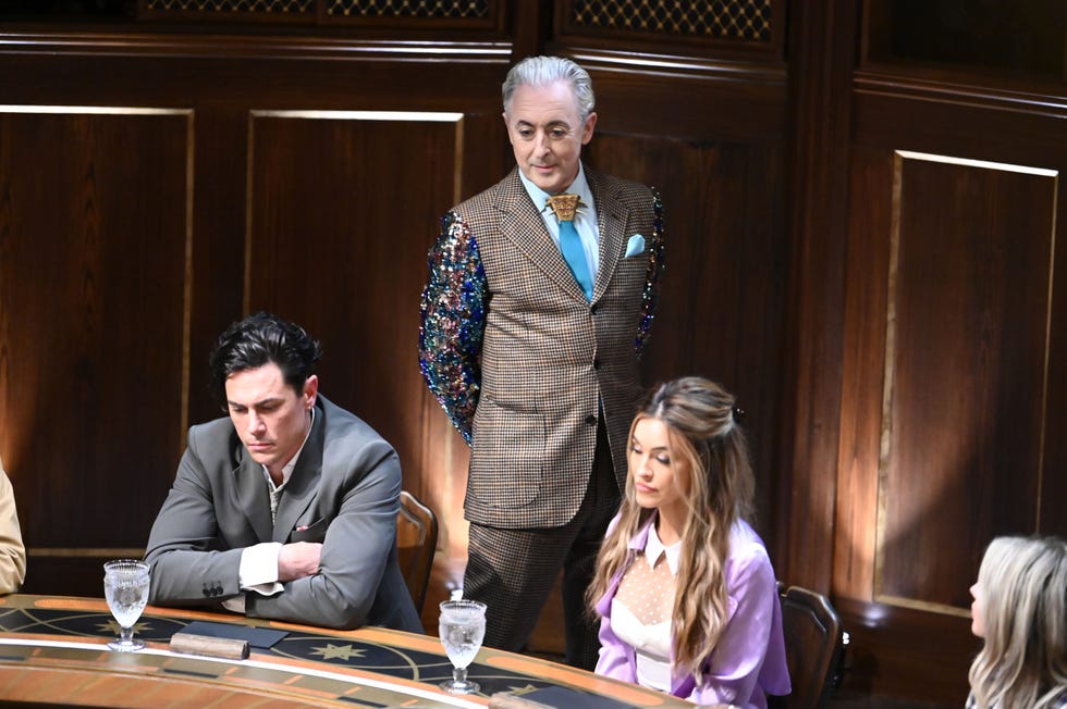 group of individuals seated at a gaming table with one standing dressed in an elaborate suit