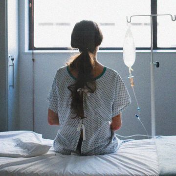 a woman sits alone in a hospital gown facing a window