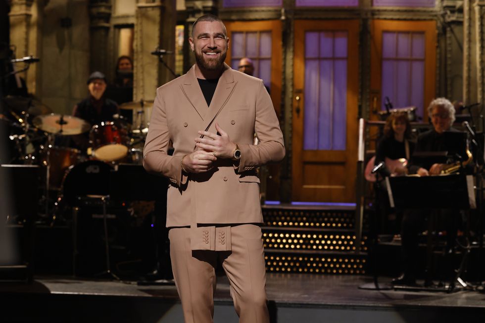 travis kelce, wearing a tan suit and pants, stands on the stage at saturday night live, smiling